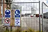 Billedet viser et hegn foran et byggeområde i Nakskov. I baggrunden ses en høj bygning indhyllet i plastik, og på hegnet ses advarselsskilte med overvågning og påbud om at bære sikkerhedsudstyr.